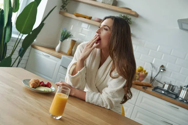 Modern mutfakta kahvaltı ederken bornozlu çekici genç bir kadın esniyor.