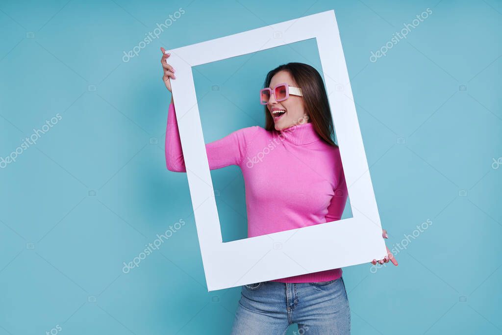 Playful young woman looking through a picture frame while standing against blue background