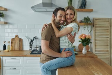 Mutfak tezgahında oturan kadın kameraya bakarken mutlu genç çift kaynaşıyor.