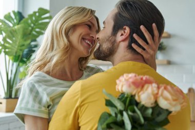 Yakışıklı genç adam mutfakta bir demet çiçekle kız arkadaşına sürpriz yapıyor.