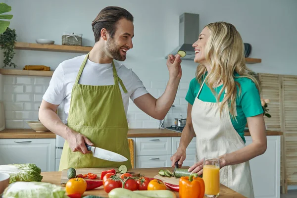 Cheerful couple feeding each other with fresh veggies while cooking at the kitchen together