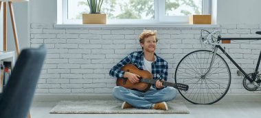 Evde yerde otururken akustik gitar çalan yakışıklı kızıl saçlı adam.
