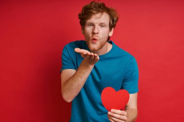 Kızıl saçlı yakışıklı adam kırmızı arka planda dururken kırmızı kağıt tutuyor ve öpücük yolluyor.