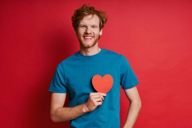 Kırmızı arka planda dikilirken kırmızı kağıt kalp tutan neşeli kızıl adam.