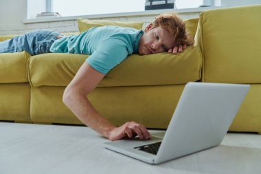 Kanepede yatarken dizüstü bilgisayar kullanan yakışıklı kızıl adam.