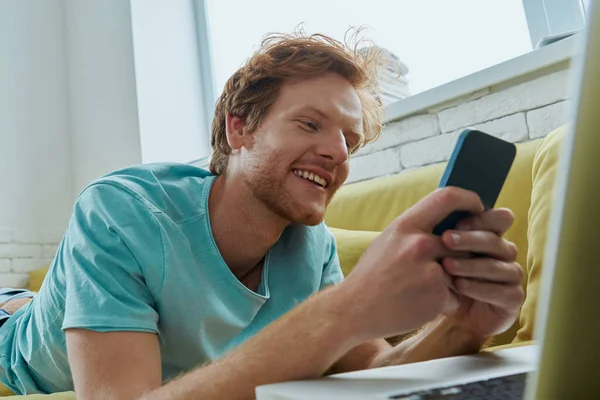Bonito Ruivo Homem Usando Tecnologias Sorrindo Enquanto Deitado Sofá Casa — Fotografia de Stock