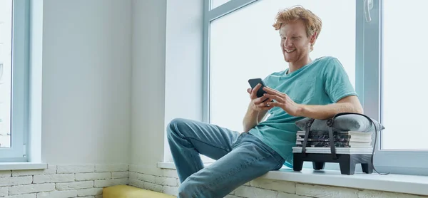 Glada Rödhårig Man Som Använder Smarttelefon När Sitter Fönsterbrädan Hemma — Stockfoto