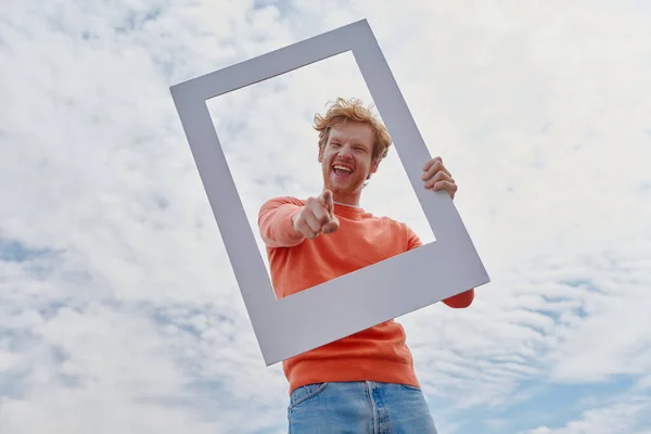 Playful young redhead man looking through a picture frame and gesturing while standing outdoors