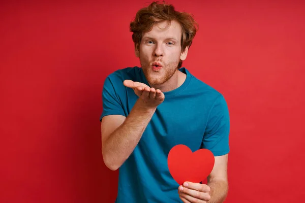 Handsome redhead man holding red paper and blowing a kiss while standing against red background