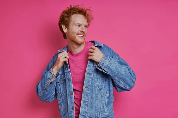 Cheerful redhead man in denim clothing standing against pink background