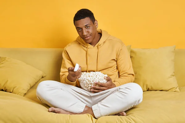 黄色の背景とソファの上でリラックスしながら テレビを見て 軽食を持っている退屈男 — ストック写真