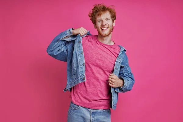Happy redhead man in denim clothing looking at camera while standing against pink background