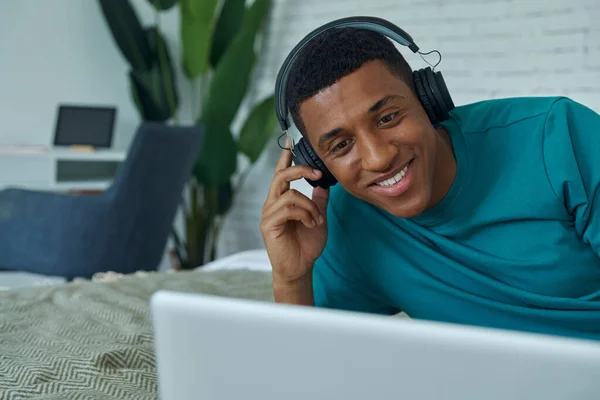 Szczęśliwy Mieszane Wyścig Człowiek Słuchawki Pomocą Laptopa Podczas Leżenia Łóżku — Zdjęcie stockowe