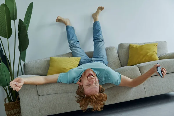 Podekscytowany Rudzielec Trzyma Smartfon Gestykuluje Relaksując Się Kanapie Domu — Zdjęcie stockowe