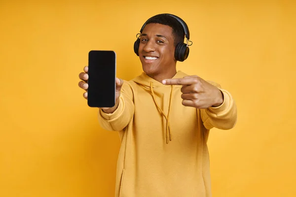 Wesoły Mieszany Człowiek Wyścig Słuchawkach Pokazuje Swój Smartfon Stojąc Przeciwko — Zdjęcie stockowe