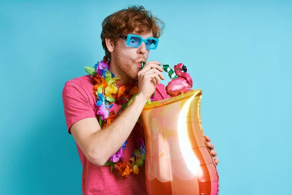 Playful man in Hawaiian necklace carrying cocktail shaped balloon against blue background