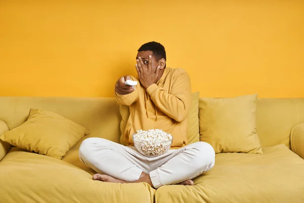 Młody Człowiek Ogląda Telewizję Wygląda Przerażony Siedząc Kanapie Żółtym Tłem — Zdjęcie stockowe