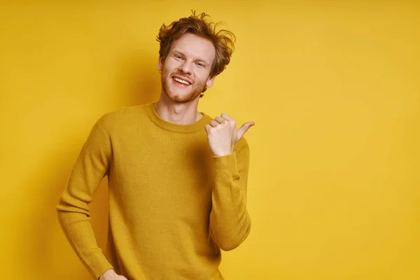 Young redhead man pointing away and smiling while standing against yellow background