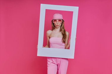 Pembe arka planda dikilirken resim çerçevesine bakan oyuncu genç kadın.