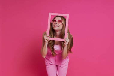 Çekici genç bir kadın pembe arka planda dikilirken bir resim çerçevesinden bakıyor.