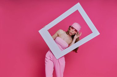 Pembe arka planda dikilirken resim çerçevesine bakan oyuncu genç kadın.