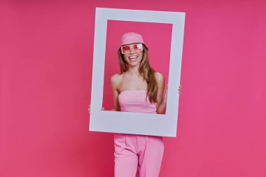 Mutlu genç bir kadın pembe arka planda dikilirken bir resim çerçevesine bakıyor.