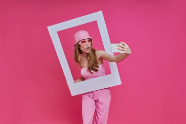 Oyuncu kadın özçekim yapıyor ve pembe arka planda dururken resim çerçevesinden bakıyor.