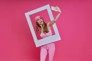 Oyuncu genç bir kadın pembe arka planda dururken özçekim yapıyor ve resim çerçevesine bakıyor.