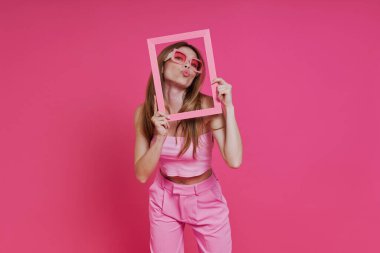 Oyuncu genç bir kadın pembe arka planda dururken resim çerçevesinden öpücük yolluyor.