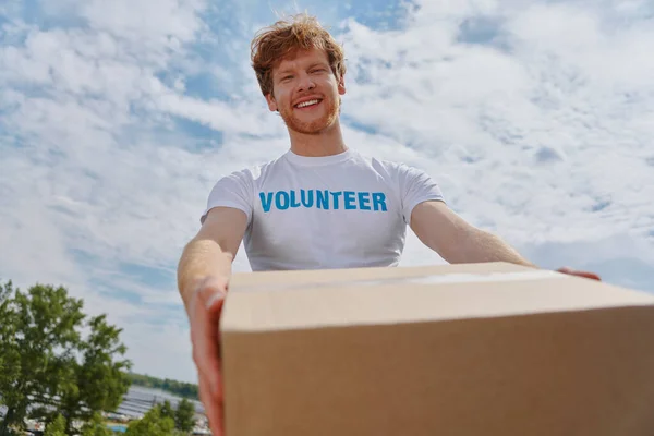 Happy young man in volunteer shirt stretching out donation box and smiling