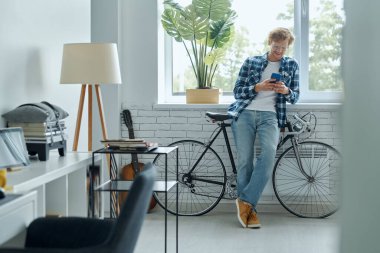 Oturma odasında bisikletinin yanında dururken akıllı telefon kullanan neşeli genç adam.