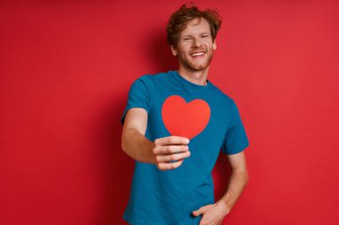 Kırmızı arka planda dikilirken kırmızı kağıt kalp gösteren mutlu kızıl adam.