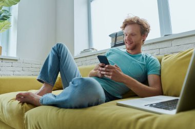 Yakışıklı kızıl saçlı adam akıllı telefon kullanıyor ve evde kanepede otururken gülümsüyor.