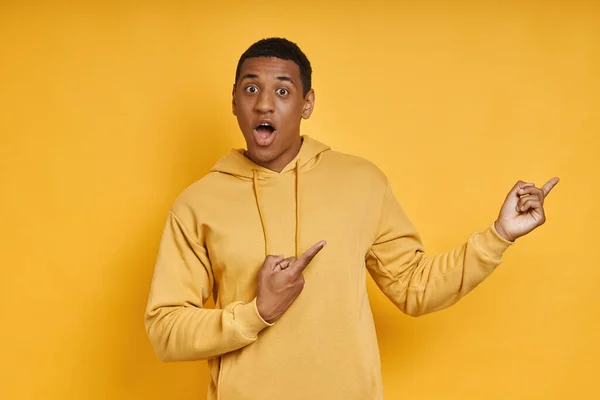 Surprised mixed race man pointing away while standing against yellow background