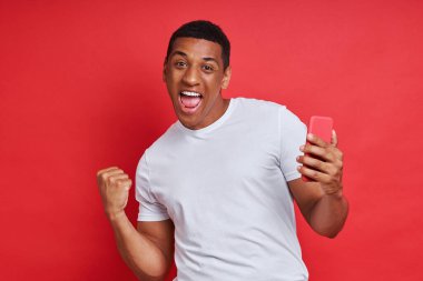 Akıllı telefon tutan ve kırmızı arka planda el kol hareketi yapan mutlu melez adam.