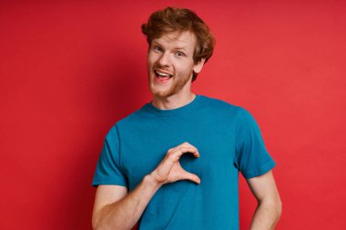 Yakışıklı kızıl saçlı adam kırmızı arka planda dikilirken yarım kalp şeklinde el kol hareketi yapıyor.