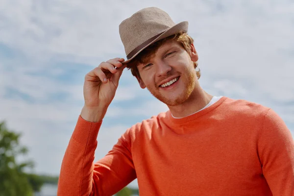 リラックスした若い赤毛の男彼の帽子と笑顔を調整しながら 屋外に立って — ストック写真