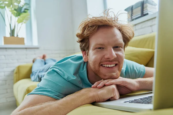 Cheerful redhead man using laptop and smiling while lying on the couch at home