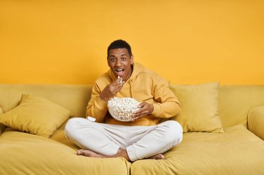 Sarı arka planda kanepede dinlenirken abur cubur yiyen mutlu yarış adamı.