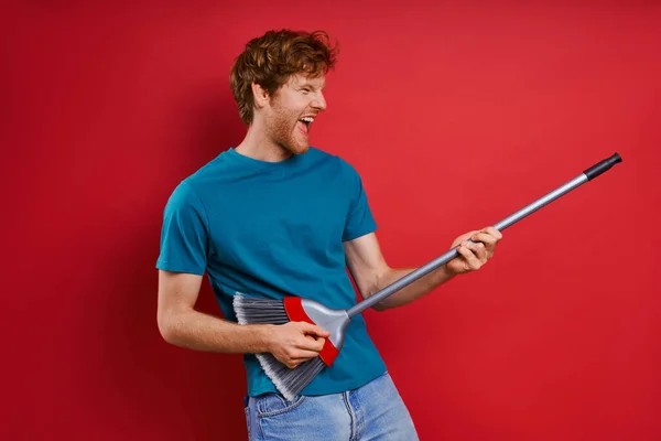 Playful Young Redhead Man Using Broom Guitar While Red Background — Stock Photo, Image