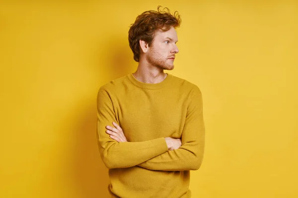 Confident redhead man keeping arms crossed while standing against yellow background