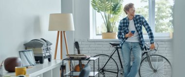 Oturma odasında bisikletinin yanında dururken cep telefonu tutan mutlu genç adam.