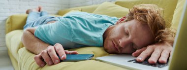 Yakışıklı kızıl saçlı adam, dizüstü bilgisayarı ve yanında akıllı telefonuyla kanepede kestiriyor.