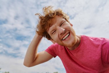 Neşeli genç kızıl adam kameraya bakıyor ve arka planda gökyüzü ile gülümsüyor.