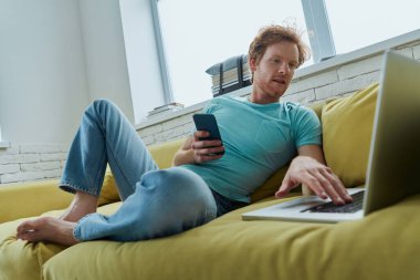Yakışıklı kızıl adam evde kanepede otururken teknoloji kullanarak akıllı davranıyor.