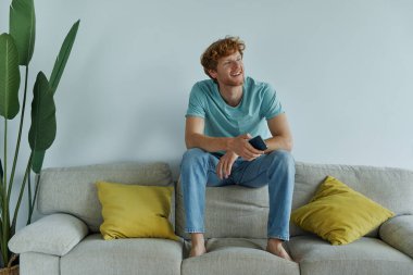 Yakışıklı kızıl saçlı adam evde kanepede dinlenirken akıllı bir telefon tutuyor.