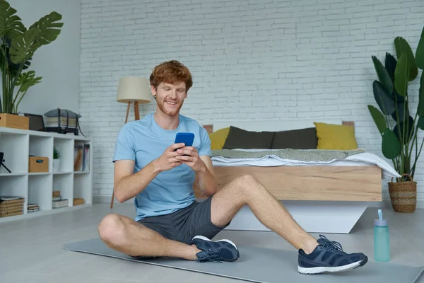 Stilig Man Sportkläder Med Smarttelefon Sittande Träningsmatta Hemma — Stockfoto