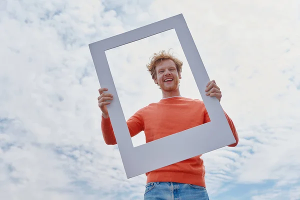 Happy young redhead man looking through a picture frame and smiling while standing outdoors