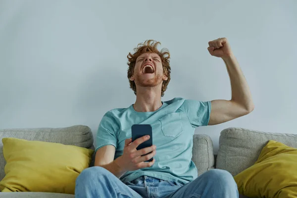 Happy Redhead Man Holding Smart Phone Gesturing While Sitting Couch — Stockfoto