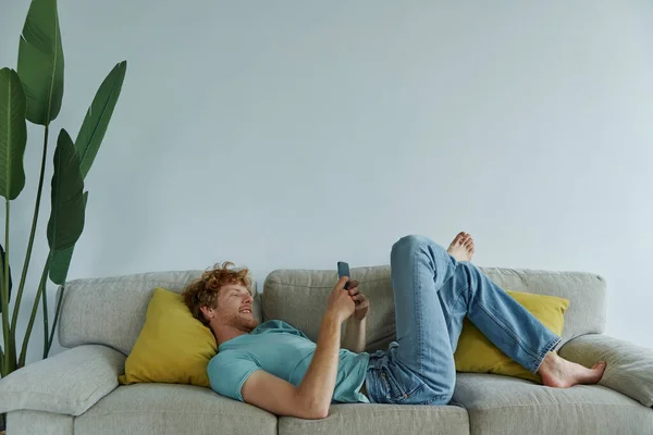 Relaxado Ruiva Homem Usando Telefone Inteligente Enquanto Deitado Sofá Casa — Fotografia de Stock
